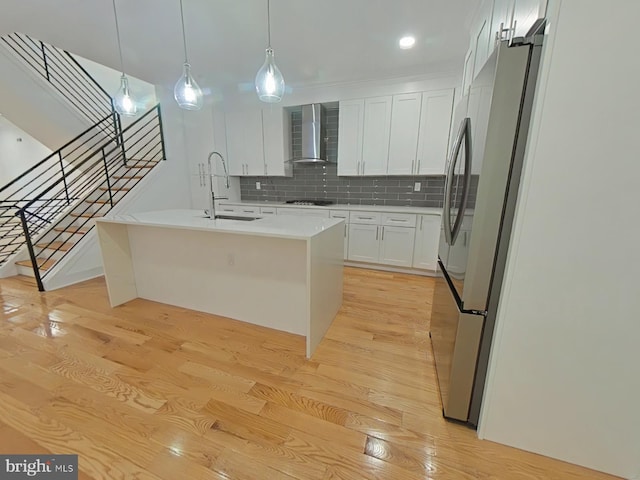 kitchen featuring sink, wall chimney range hood, stainless steel fridge, a kitchen island with sink, and light wood-type flooring