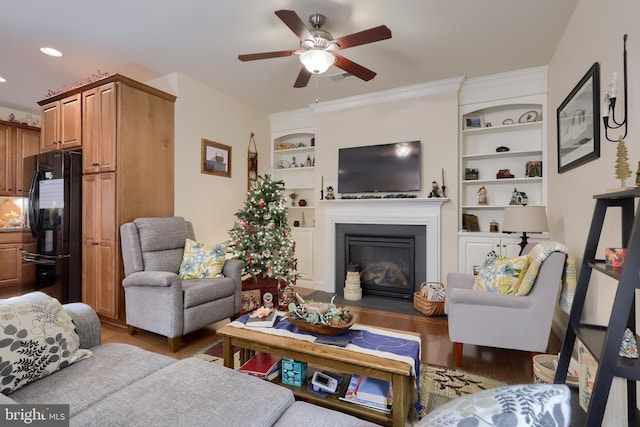 living area featuring visible vents, built in features, a ceiling fan, a glass covered fireplace, and wood finished floors