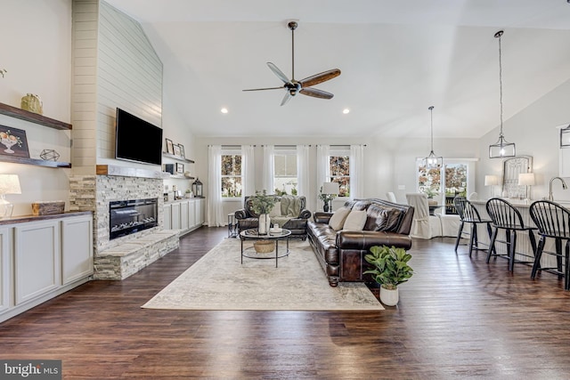 living room with ceiling fan with notable chandelier, a stone fireplace, dark wood-type flooring, and high vaulted ceiling