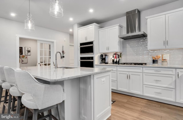 kitchen with a center island with sink, white cabinets, wall chimney range hood, sink, and light hardwood / wood-style flooring