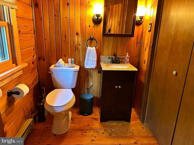 bathroom with a baseboard heating unit, wood-type flooring, toilet, wooden walls, and vanity