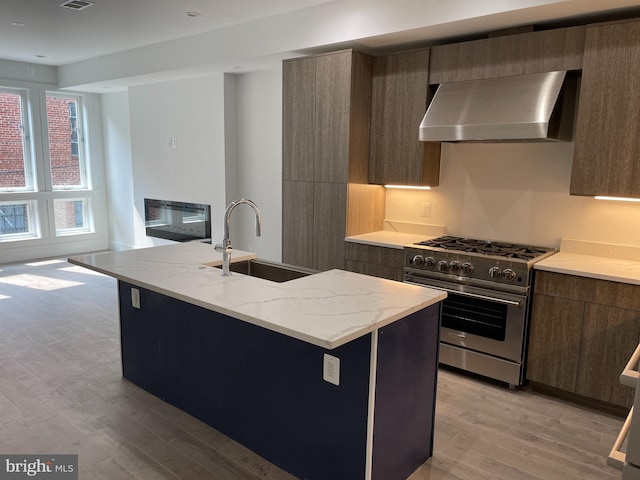 kitchen featuring sink, wall chimney exhaust hood, light hardwood / wood-style floors, high end stainless steel range, and a center island with sink