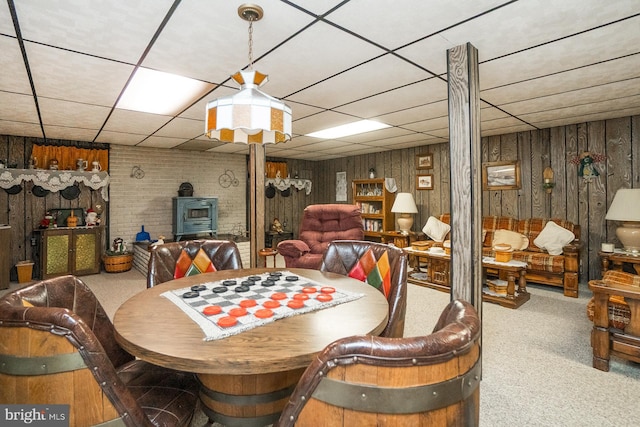 dining area with carpet floors, a wood stove, a drop ceiling, and wood walls