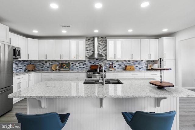 kitchen with white cabinets, wall chimney range hood, an island with sink, appliances with stainless steel finishes, and a breakfast bar area