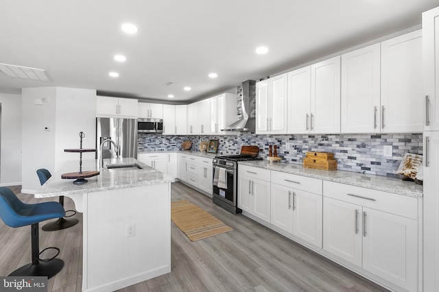 kitchen featuring light stone countertops, appliances with stainless steel finishes, wall chimney range hood, light hardwood / wood-style flooring, and white cabinets