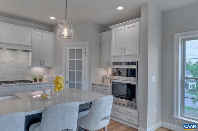 kitchen with light stone countertops, appliances with stainless steel finishes, ventilation hood, white cabinetry, and hanging light fixtures