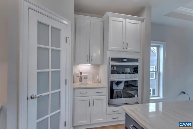 kitchen with white cabinets, light stone counters, stainless steel double oven, and backsplash