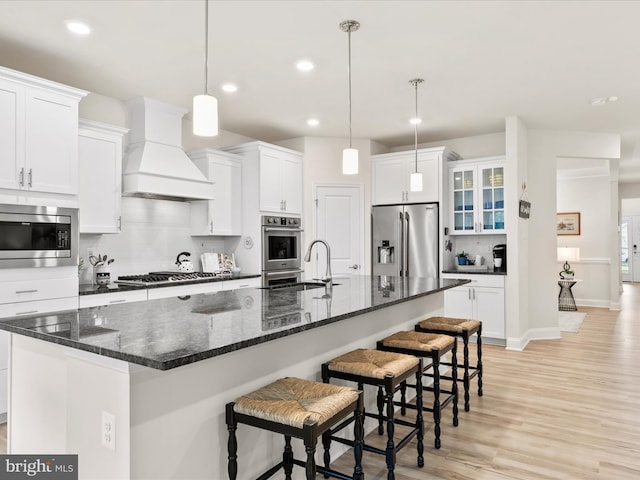 kitchen featuring stainless steel appliances, premium range hood, pendant lighting, a center island with sink, and white cabinets