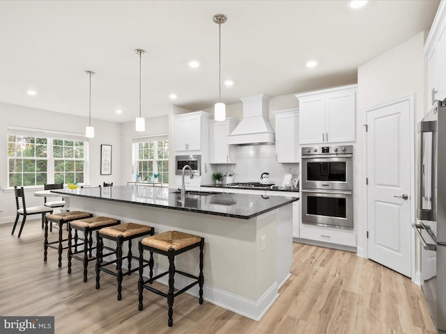 kitchen with appliances with stainless steel finishes, white cabinets, premium range hood, and a kitchen island with sink