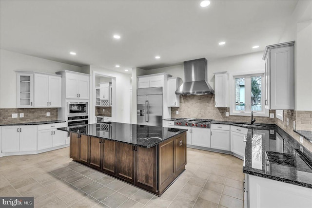 kitchen featuring a center island, sink, wall chimney range hood, built in appliances, and dark stone countertops