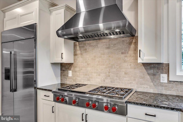 kitchen with appliances with stainless steel finishes, tasteful backsplash, dark stone counters, wall chimney range hood, and white cabinets