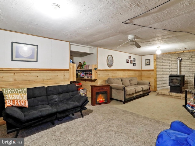 carpeted living room with ceiling fan, a wood stove, a textured ceiling, and wooden walls