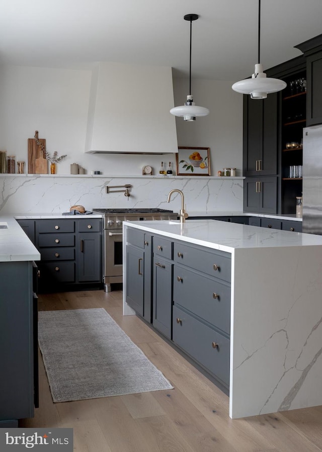 kitchen with light wood-type flooring, light stone counters, exhaust hood, pendant lighting, and a center island with sink