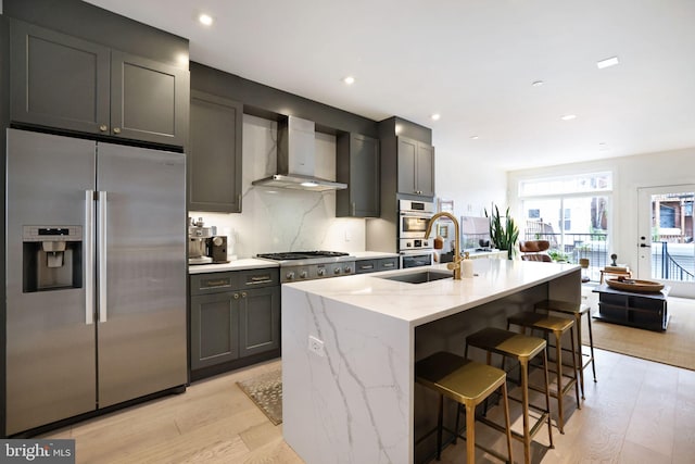 kitchen featuring sink, stainless steel appliances, wall chimney range hood, light hardwood / wood-style flooring, and a center island with sink