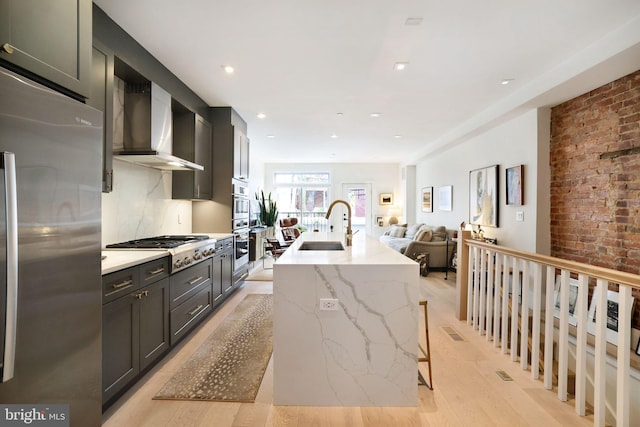 kitchen with sink, wall chimney exhaust hood, light hardwood / wood-style floors, a center island with sink, and appliances with stainless steel finishes