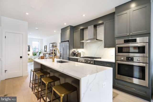 kitchen featuring appliances with stainless steel finishes, a center island with sink, wall chimney exhaust hood, and gray cabinetry