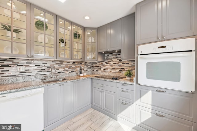 kitchen featuring tasteful backsplash, gray cabinets, glass insert cabinets, a sink, and white appliances