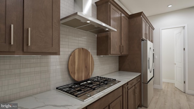 kitchen with white fridge with ice dispenser, wall chimney range hood, light stone counters, light hardwood / wood-style flooring, and stainless steel gas stovetop