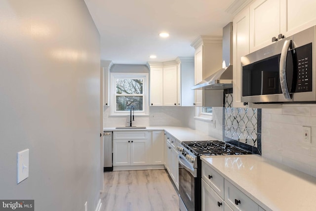 kitchen featuring wall chimney range hood, sink, appliances with stainless steel finishes, light hardwood / wood-style floors, and white cabinetry