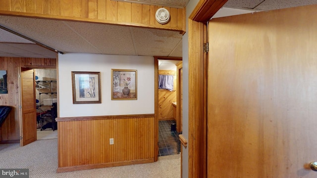 hallway with a paneled ceiling, wood walls, and light carpet