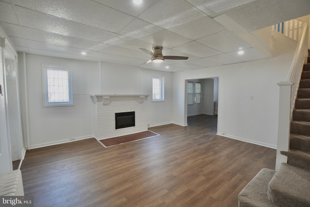 unfurnished living room featuring ceiling fan, a drop ceiling, dark hardwood / wood-style flooring, and a brick fireplace