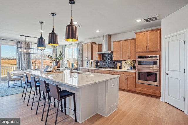 kitchen with light stone counters, decorative light fixtures, appliances with stainless steel finishes, an island with sink, and wall chimney range hood