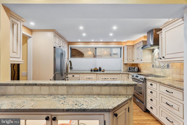 kitchen with cream cabinetry, appliances with stainless steel finishes, light stone countertops, and wall chimney range hood