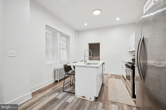 kitchen with radiator heating unit, sink, stainless steel appliances, a kitchen island with sink, and white cabinets