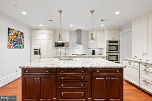 kitchen featuring stainless steel appliances, wall chimney exhaust hood, decorative light fixtures, and a kitchen island with sink