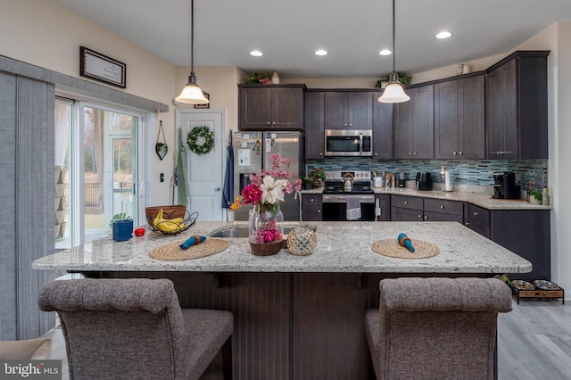 kitchen with appliances with stainless steel finishes, decorative light fixtures, light stone counters, and a center island with sink