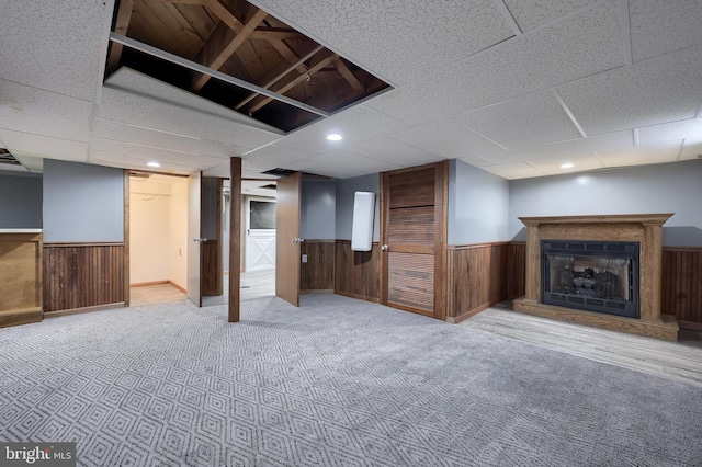 basement with a drop ceiling, wood walls, and light colored carpet