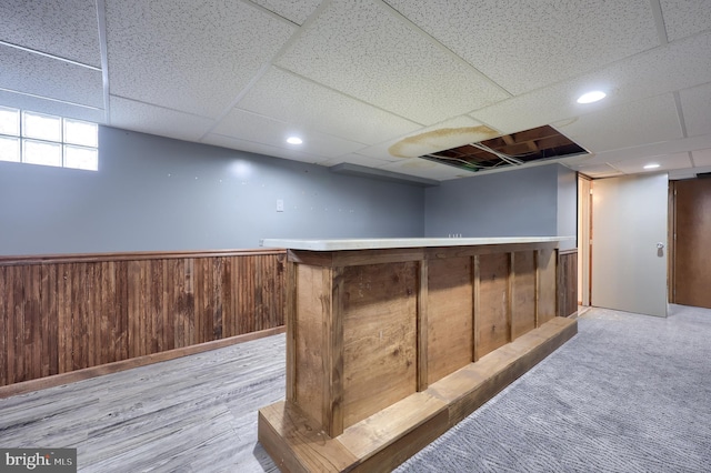 basement featuring a drop ceiling, wooden walls, indoor bar, and light wood-type flooring