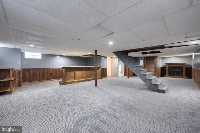 basement featuring a paneled ceiling, light carpet, wood walls, and bar area