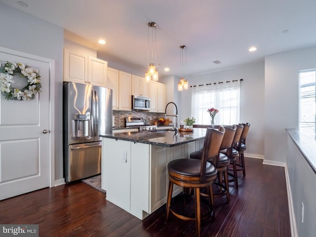 kitchen with a kitchen island with sink, dark stone counters, appliances with stainless steel finishes, a healthy amount of sunlight, and dark hardwood / wood-style flooring