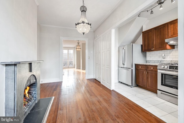 kitchen with a warm lit fireplace, light wood-style flooring, under cabinet range hood, stainless steel appliances, and backsplash