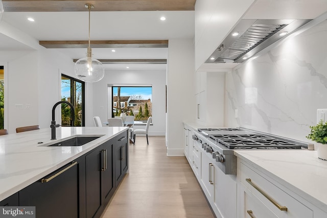 kitchen with wall chimney range hood, sink, pendant lighting, beam ceiling, and white cabinets