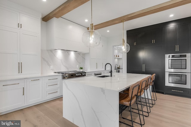 kitchen with white cabinetry, sink, hanging light fixtures, beamed ceiling, and an island with sink