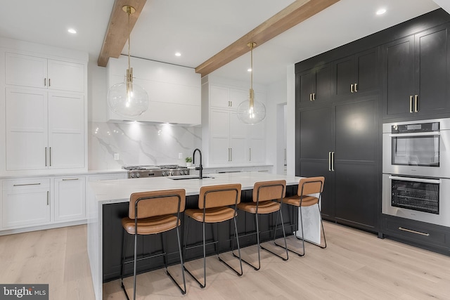 kitchen featuring double oven, beam ceiling, pendant lighting, a center island with sink, and white cabinetry