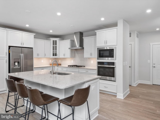 kitchen featuring sink, wall chimney range hood, stainless steel appliances, a kitchen island with sink, and white cabinets