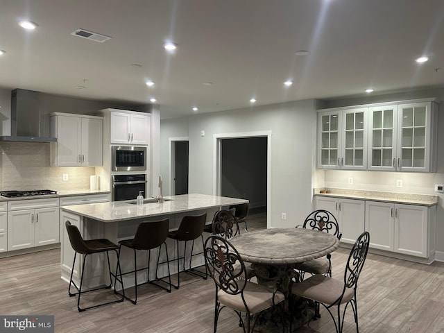 kitchen with a sink, appliances with stainless steel finishes, wall chimney exhaust hood, light wood finished floors, and a center island with sink