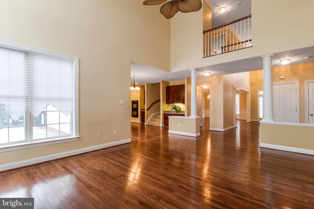 unfurnished living room featuring ceiling fan with notable chandelier, a high ceiling, dark hardwood / wood-style flooring, and ornate columns