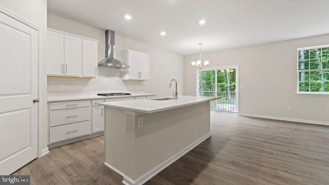 kitchen with wall chimney range hood, an island with sink, white cabinetry, and sink