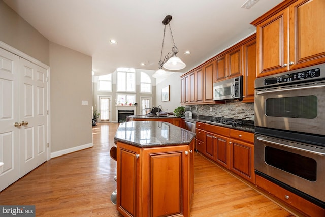 kitchen featuring appliances with stainless steel finishes, light wood-type flooring, decorative light fixtures, dark stone countertops, and a center island