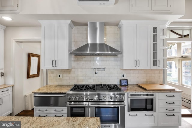 kitchen featuring backsplash, white cabinets, wall chimney exhaust hood, light stone countertops, and appliances with stainless steel finishes