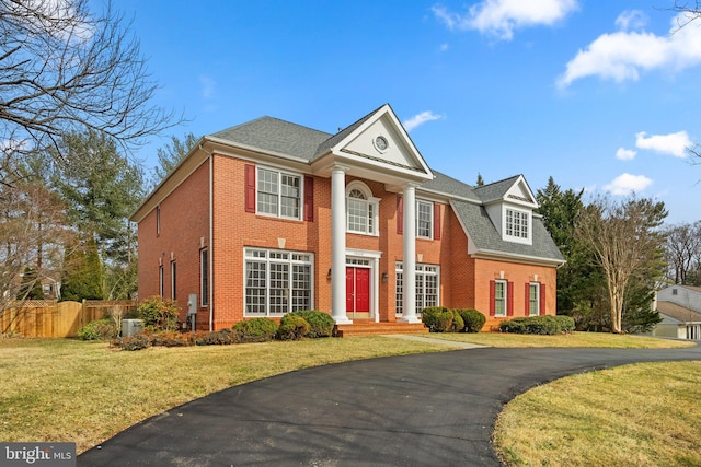 neoclassical / greek revival house featuring brick siding, roof with shingles, a front yard, and fence