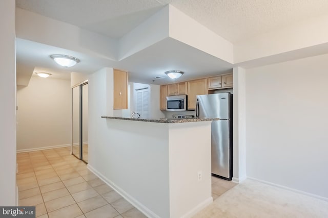 kitchen with dark stone countertops, light tile patterned floors, light brown cabinetry, appliances with stainless steel finishes, and kitchen peninsula