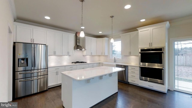 kitchen with appliances with stainless steel finishes, a kitchen island, pendant lighting, wall chimney range hood, and white cabinets