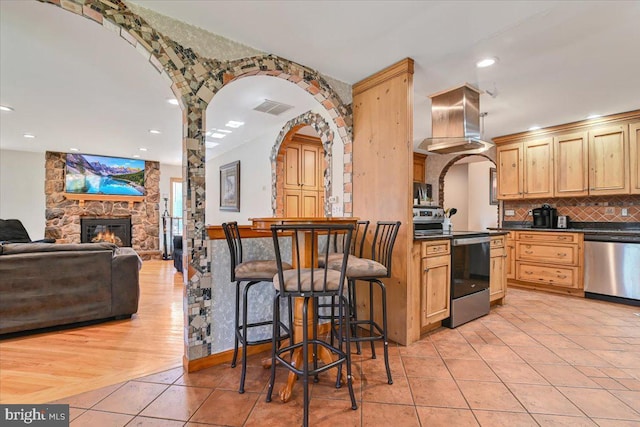 kitchen featuring stainless steel appliances, extractor fan, light brown cabinets, light hardwood / wood-style flooring, and a stone fireplace