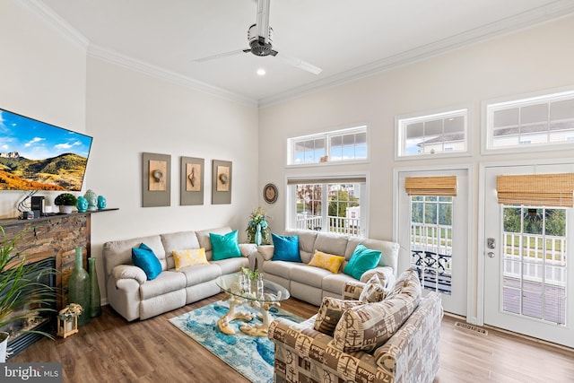 living room featuring ceiling fan, crown molding, light hardwood / wood-style flooring, a high ceiling, and a stone fireplace