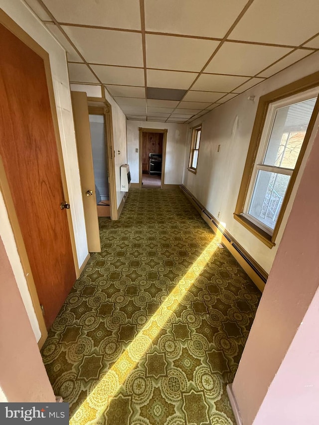 hallway featuring a drop ceiling, a baseboard heating unit, and dark colored carpet